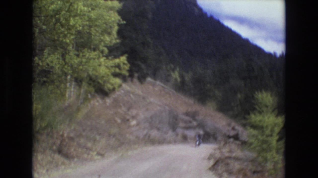In metadata-supported 1973 Colorado Springs-area footage, a small helmeted motorcyclist appears on an unpaved mountain road bordered by trees, rocks, and forested slopes.
