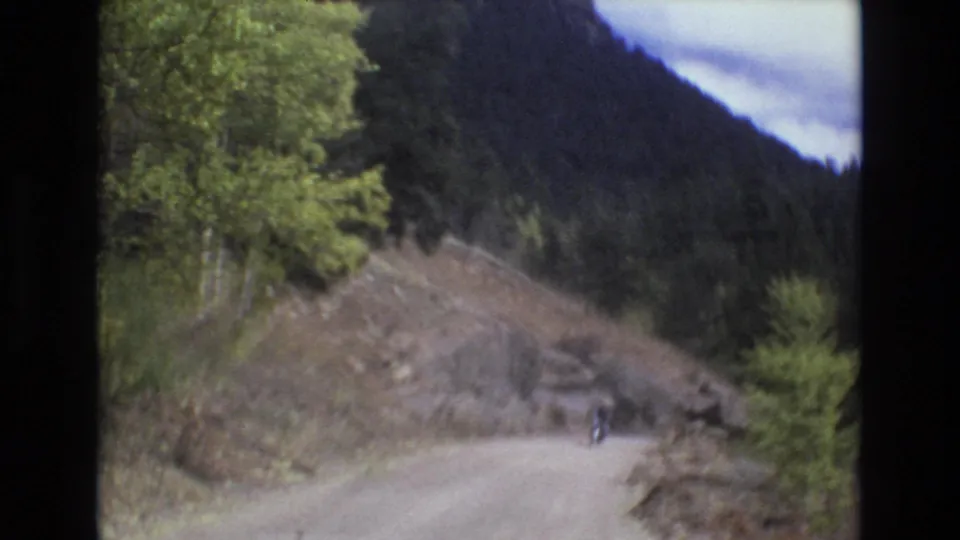In metadata-supported 1973 Colorado Springs-area footage, a small helmeted motorcyclist appears on an unpaved mountain road bordered by trees, rocks, and forested slopes.