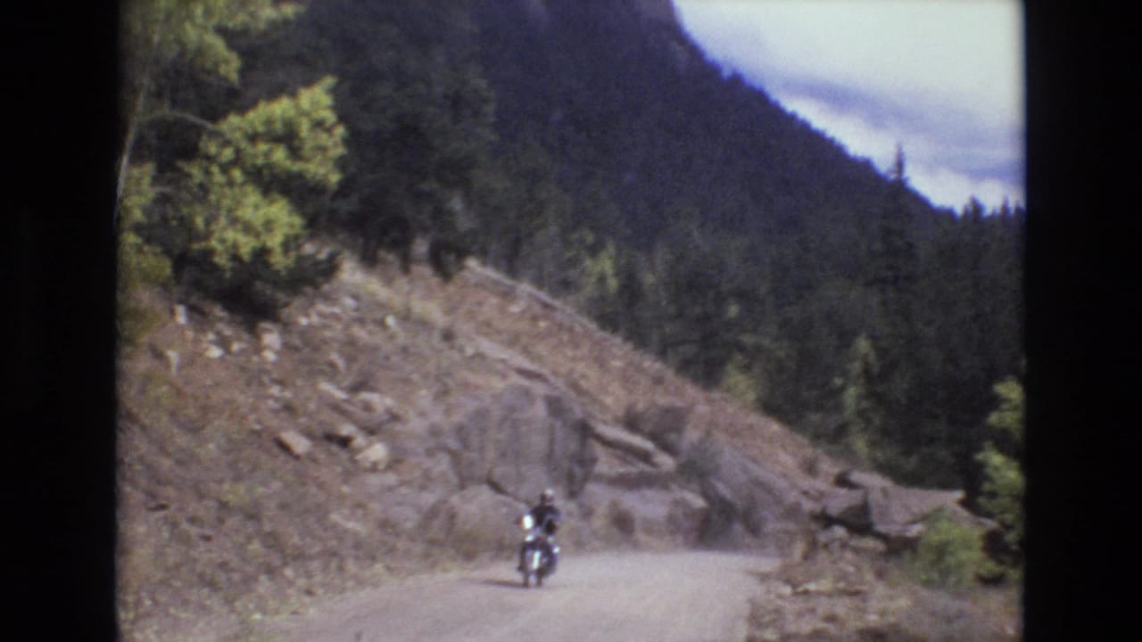 A motorcyclist rides closer on a dusty mountain road below a rocky hillside.