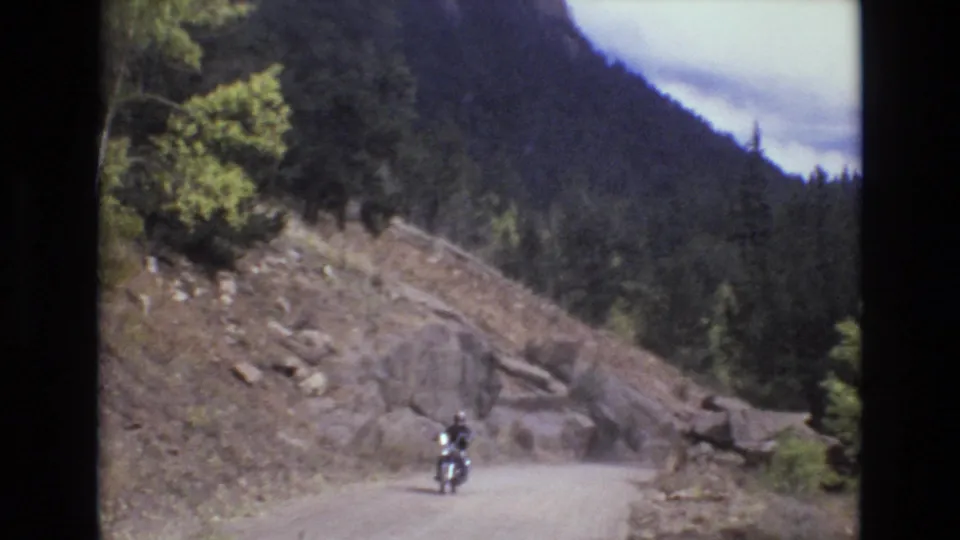 A helmeted rider moves along a dusty unpaved road in the Colorado mountain setting, with loose rock, evergreen forest, and a dark mountain ridge behind them.