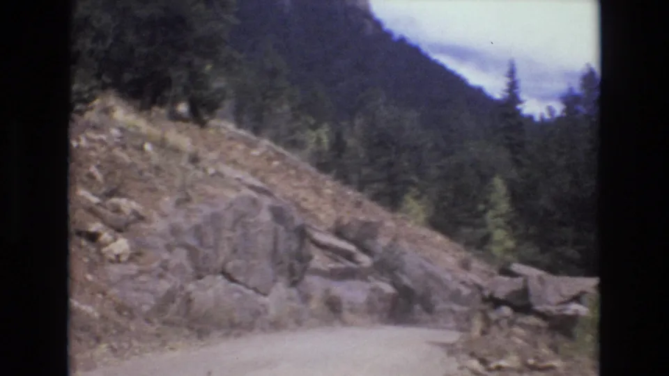 The frame shows the curving unpaved mountain road, large roadside rocks, loose gravel, trees, and distant forested slopes in the 1970s home-movie travel sequence.