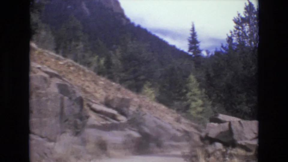 The closing view lingers on the unpaved road, roadside boulders, sloped rock cut, evergreen trees, and wooded Colorado mountain backdrop after the rider exits the frame.