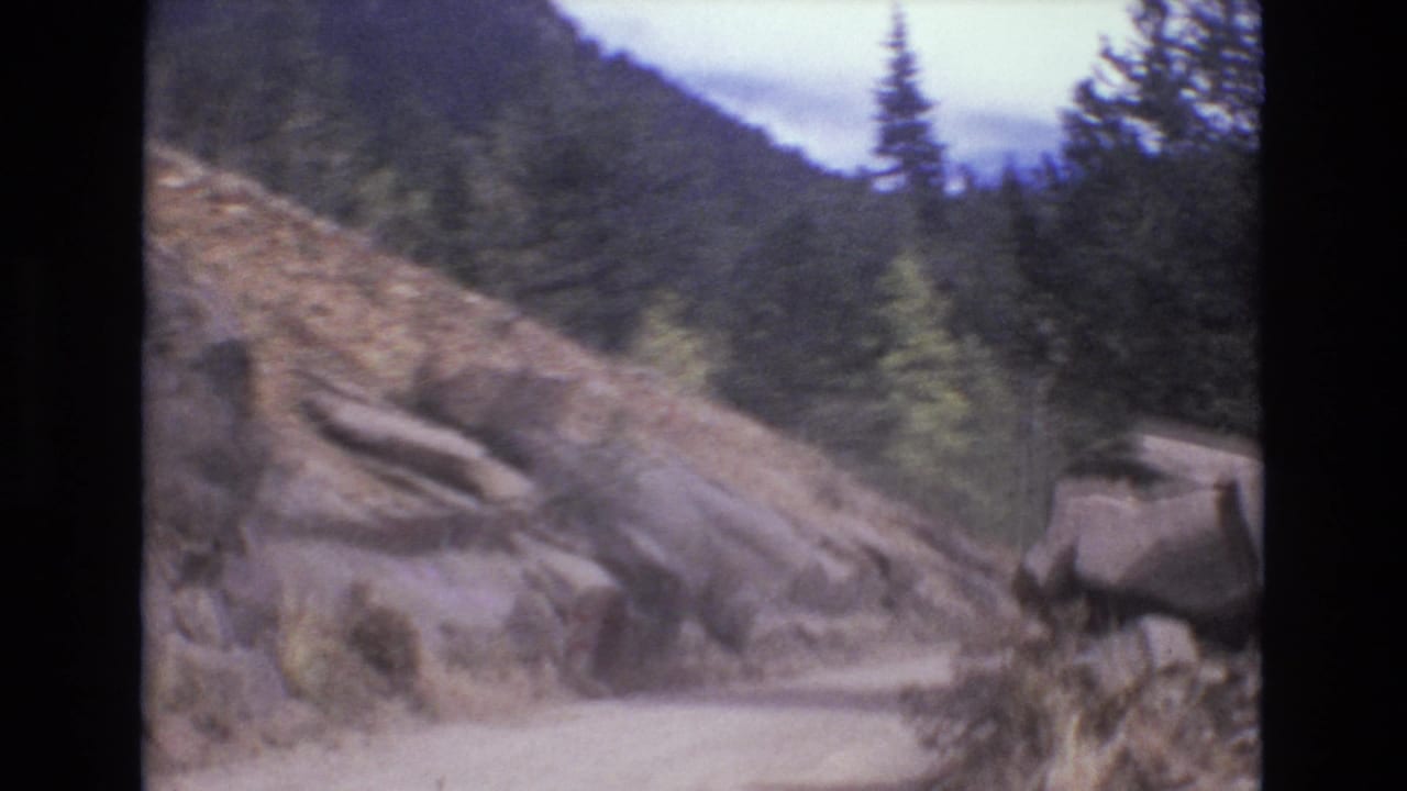 1973 8mm home-movie frame of a narrow dirt road curving past boulders, scrub, and evergreen trees in the Colorado Springs mountain area.