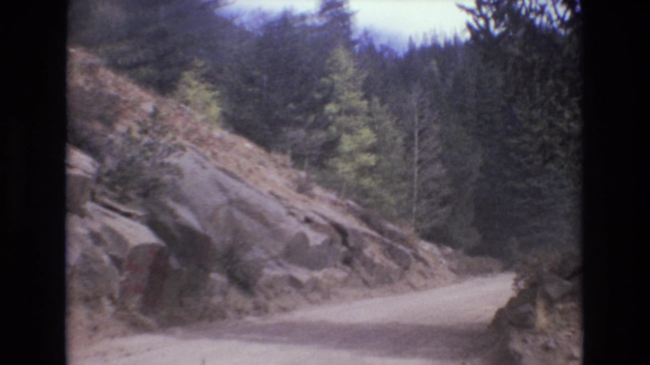 A rocky roadside bend appears in 1973 Colorado Springs 8mm footage, with pale dirt road, exposed stone, and dense evergreen forest beyond.