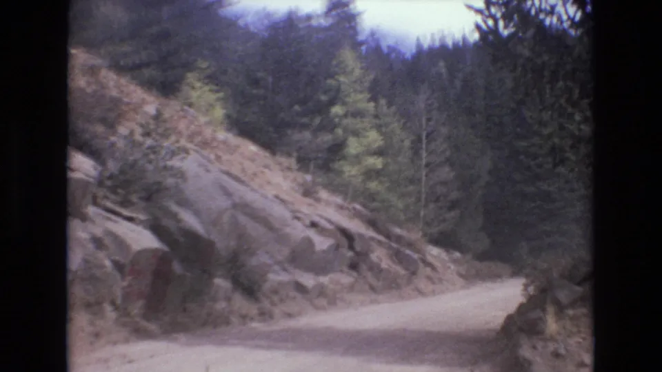 A rocky roadside bend appears in 1973 Colorado Springs 8mm footage, with pale dirt road, exposed stone, and dense evergreen forest beyond.