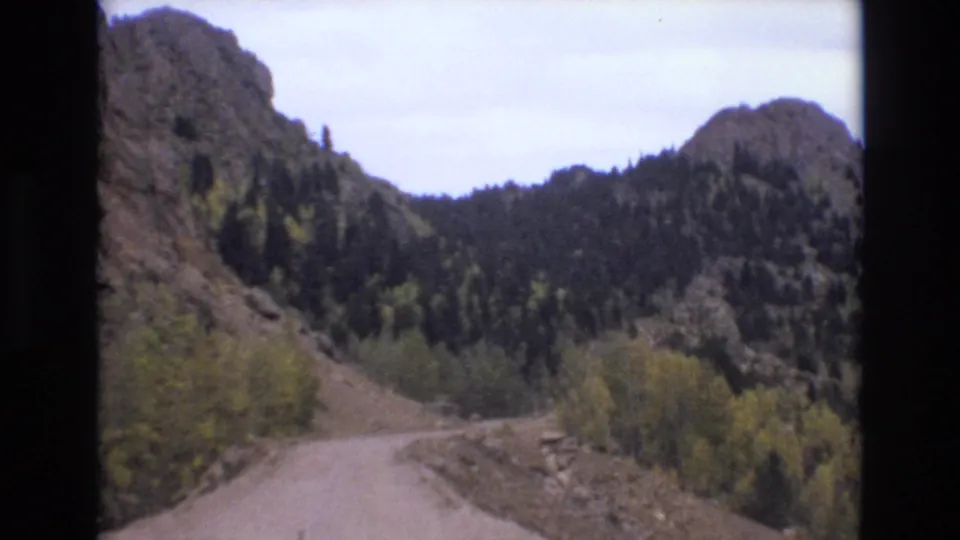 A curving dirt mountain road runs below rocky cliffs and wooded slopes in 1973 Colorado Springs home-movie footage.