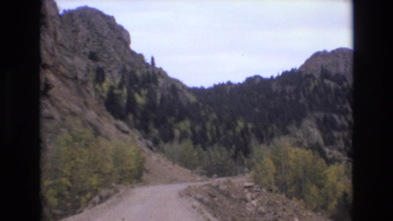 1973 8mm home-movie frame from Colorado Springs showing a dirt road curving through a rocky, forested mountain valley.