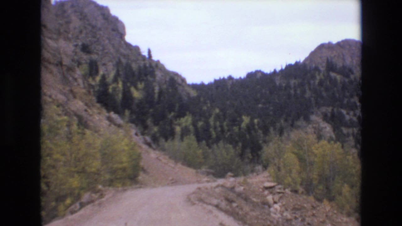 1973 Colorado Springs 8mm frame showing a curving unpaved road, rocky canyon slopes, evergreen trees, and autumn foliage.