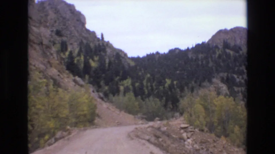 1973 Colorado Springs 8mm frame showing a curving unpaved road, rocky canyon slopes, evergreen trees, and autumn foliage.