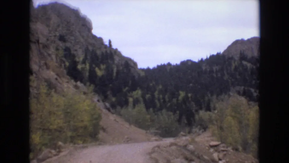 Soft 8mm frame from the Colorado Springs travel clip showing a dirt road, yellow trees, rocky hillside, and dark conifers.