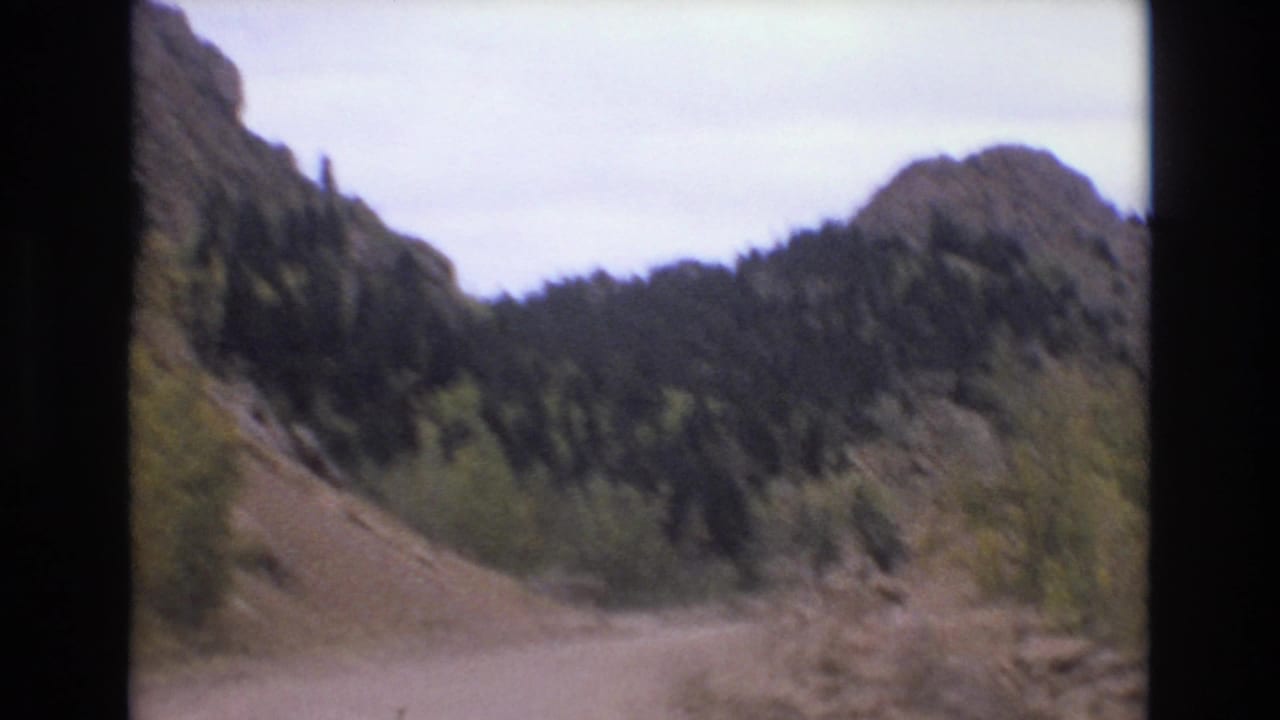 1973 Colorado Springs 8mm frame showing a dirt road entering rocky mountain terrain with evergreen forest and muted autumn color.