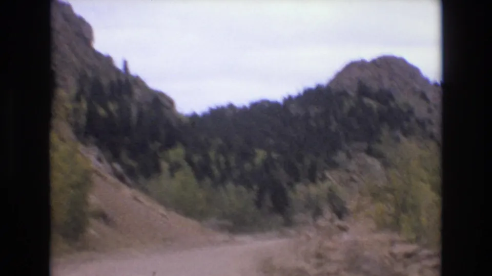 1973 Colorado Springs 8mm frame showing a dirt road entering rocky mountain terrain with evergreen forest and muted autumn color.