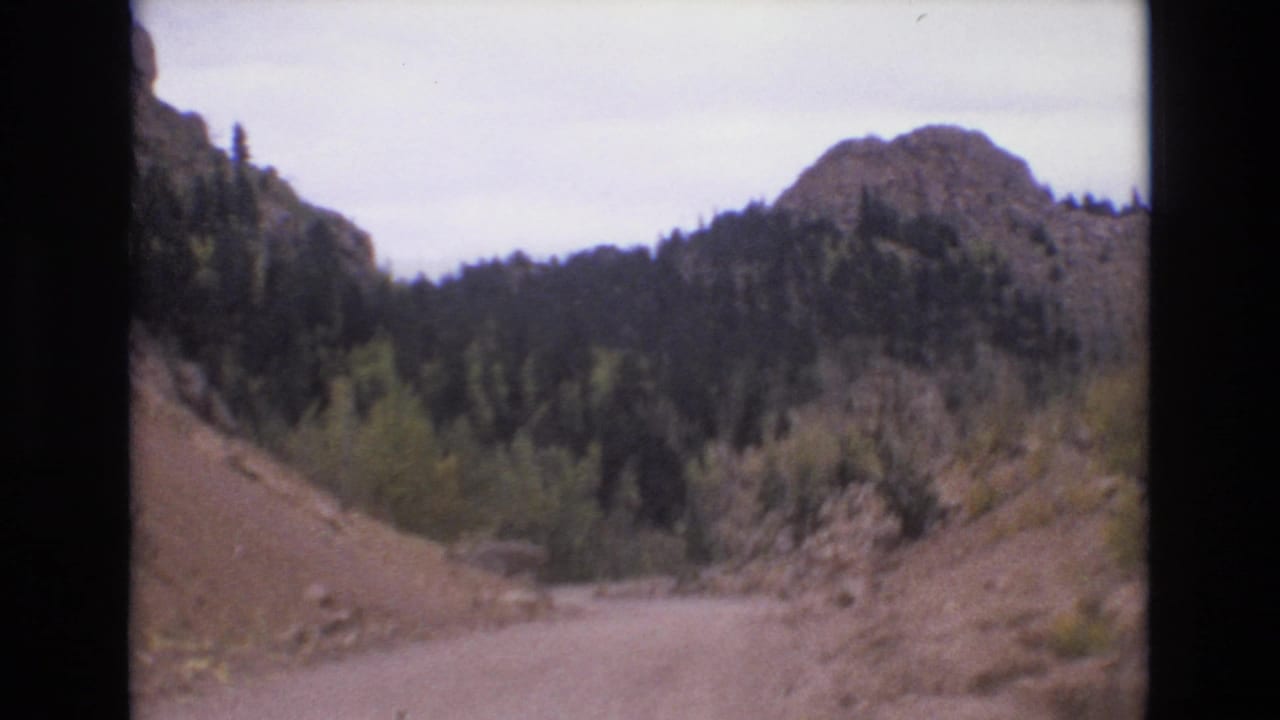 1973 Colorado Springs home-movie frame looking ahead along a winding dirt road between rocky hillsides and mixed forest.