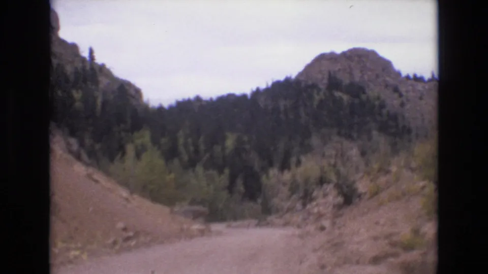 1973 Colorado Springs home-movie frame looking ahead along a winding dirt road between rocky hillsides and mixed forest.