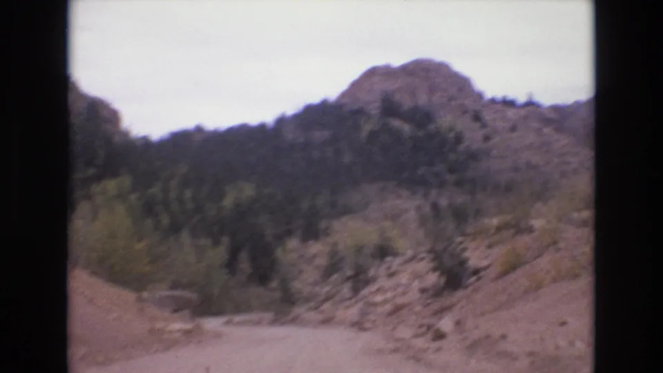 1973 Colorado Springs 8mm frame showing the road area and surrounding rocky slopes, evergreen trees, and a rounded mountain ridge.