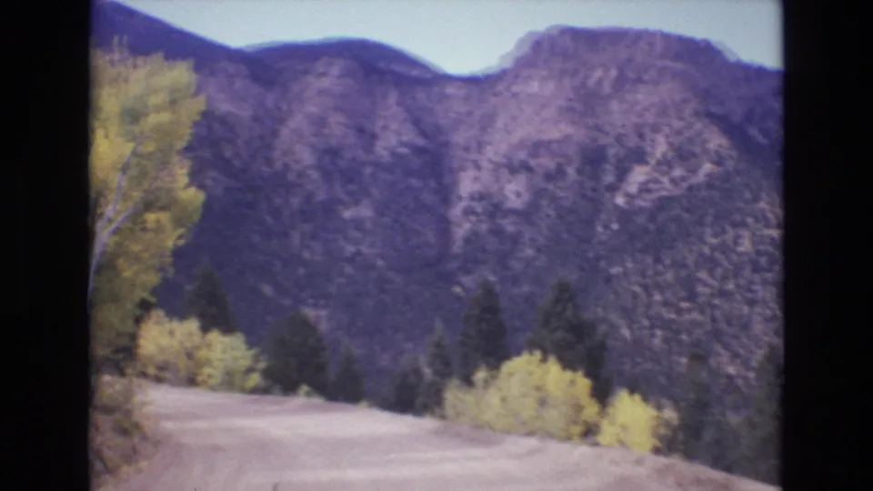 1973 Colorado Springs home-movie frame looking down a dirt mountain road with yellow autumn trees and a steep rocky ridge beyond.