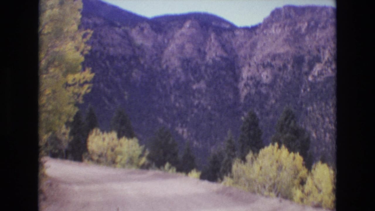 1973 Colorado Springs-area 8mm frame showing a dirt road bending through yellowing aspens and dark evergreens beneath rugged mountain slopes.