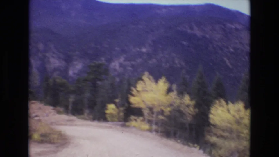 Soft 8mm frame from a moving vehicle showing a Colorado mountain road bend, yellow aspens, evergreen trees, and a dark rocky slope in the distance.