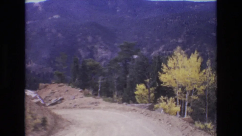 1973 Colorado Springs-area 8mm view of a dirt road curving along a forested mountainside with golden aspens, dark pines, and roadside gravel.