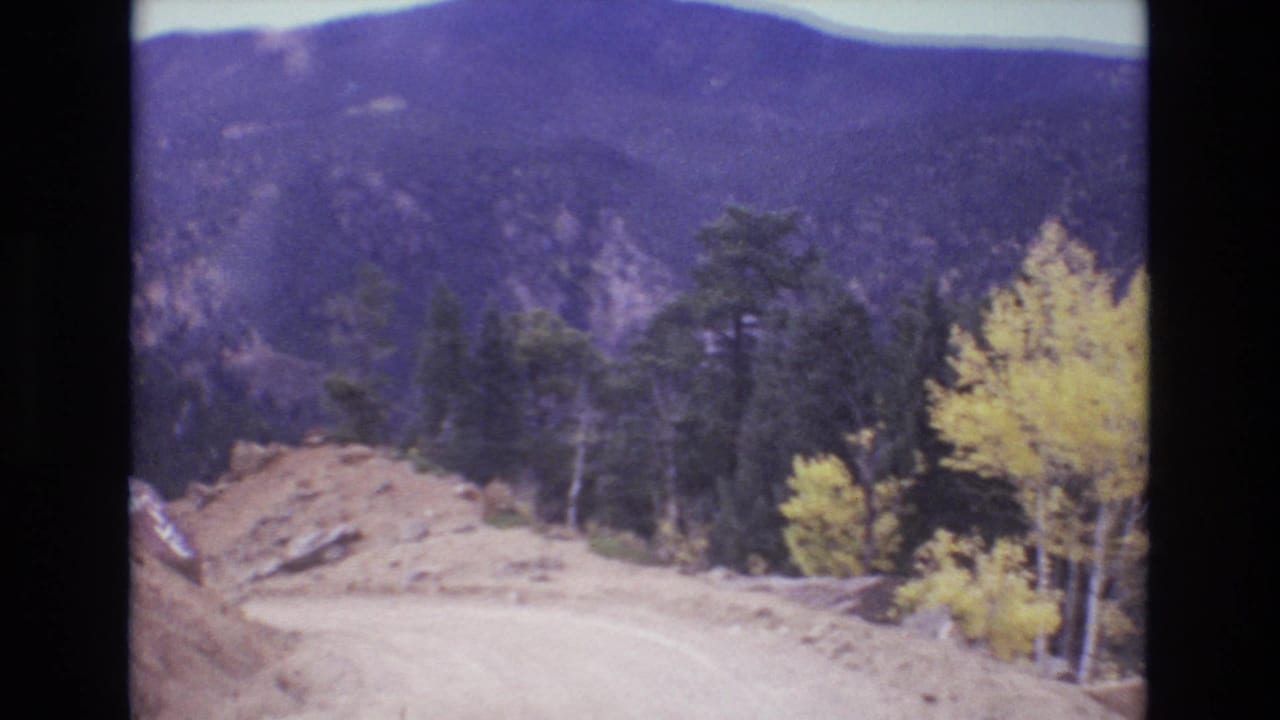 1973 Colorado Springs home-movie frame showing a dirt mountain road, a mound of loose sand or gravel, evergreen trees, yellow aspens, and distant mountain slopes.