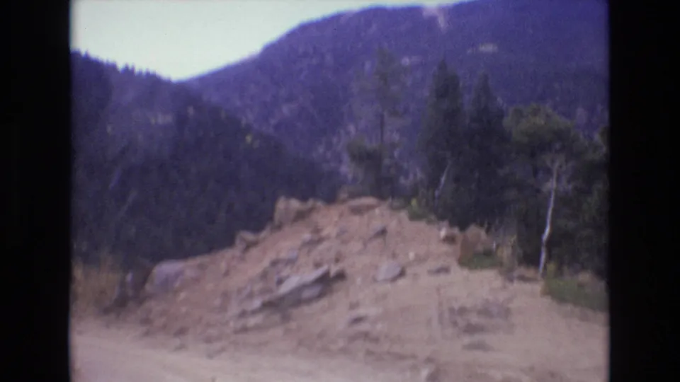 1973 Colorado Springs home-movie still showing a closer view of a rocky sand pile beside a dirt road, backed by evergreen trees and mountain slopes.