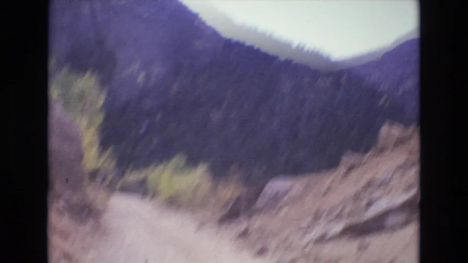 1973 Colorado Springs frame with motion blur from a moving car, looking along a dirt mountain road past a rocky roadside bank, trees, and distant slopes.