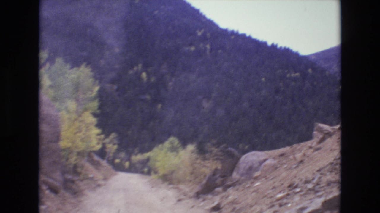 1973 Colorado Springs area 8mm frame showing a narrow dirt road ahead, rocky roadside ground, scattered boulders, yellowing trees, and a dark forested mountainside.