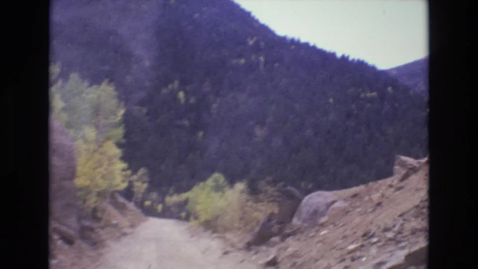 1973 Colorado Springs area 8mm frame showing a narrow dirt road ahead, rocky roadside ground, scattered boulders, yellowing trees, and a dark forested mountainside.