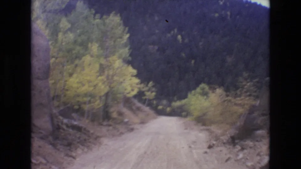 1973 Colorado Springs area 8mm frame with an unpaved road bending past yellow-green roadside trees, loose rocks, and a forested mountain backdrop.