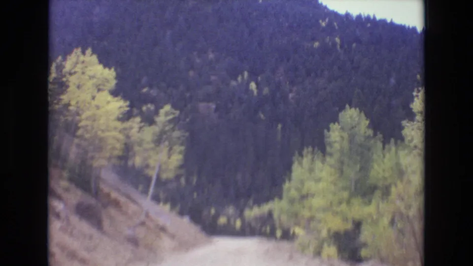 1973 Colorado Springs area 8mm frame looking forward along a dirt road bordered by yellowing aspens, green shrubs, rocky ground, and a dense evergreen mountainside.