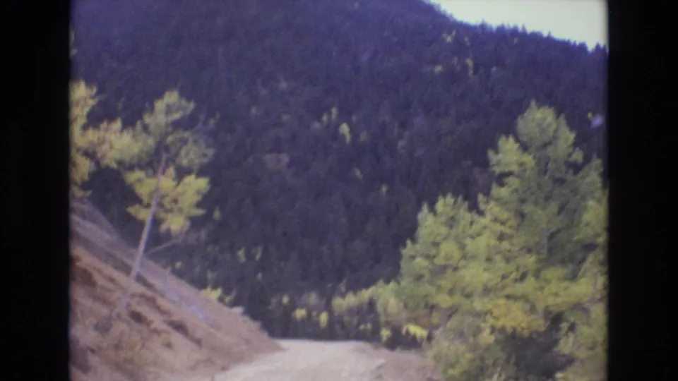 1973 Colorado Springs area 8mm frame showing the road curving past a rocky bank and autumn-colored trees, with a dark wooded mountain filling the background.