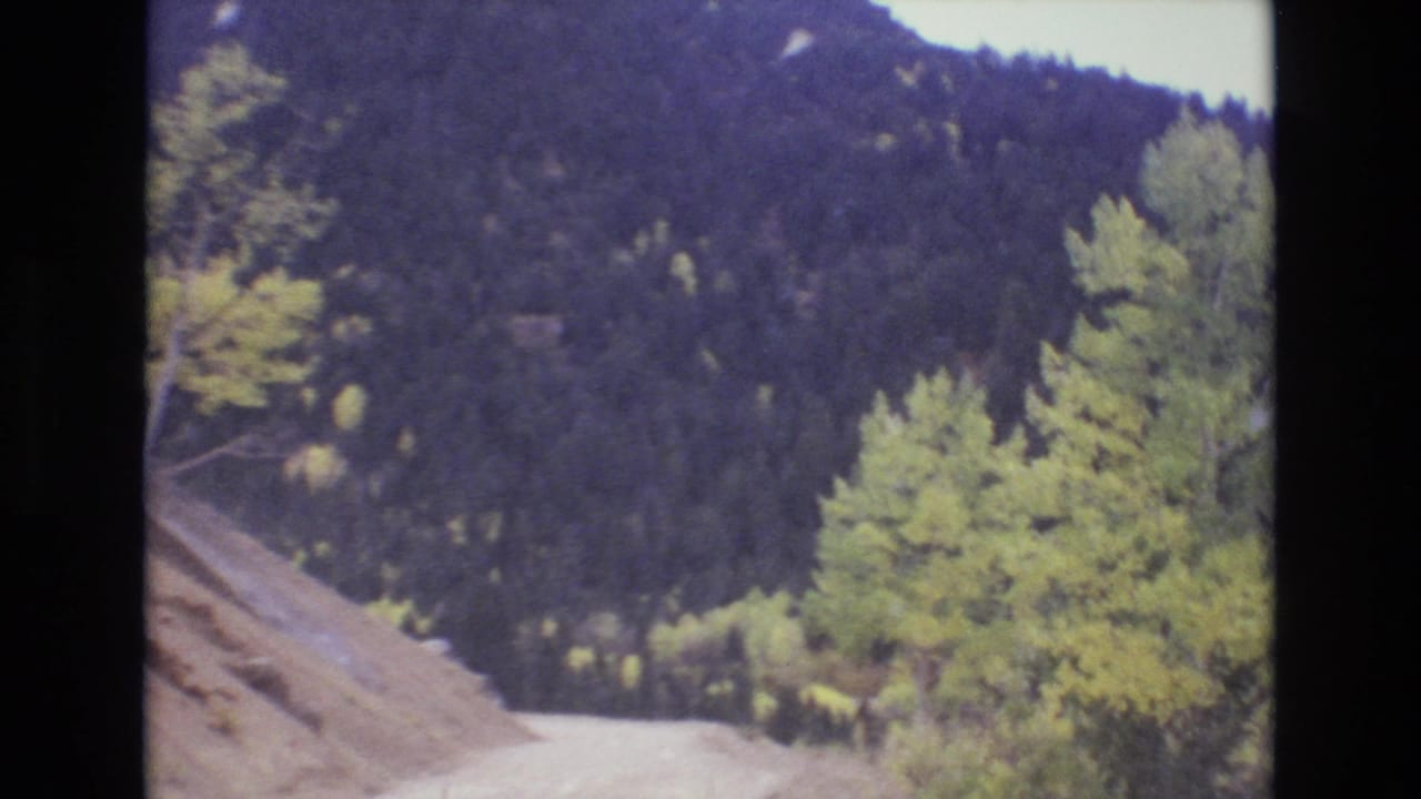 1973 Colorado Springs home-movie frame showing a pale dirt road below dark forested hills and roadside trees.