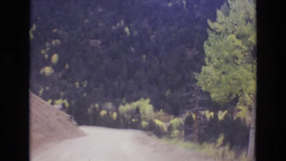 1973 Colorado Springs 8mm frame looking down an unpaved mountain road with trees and wooded slopes rising beyond it.