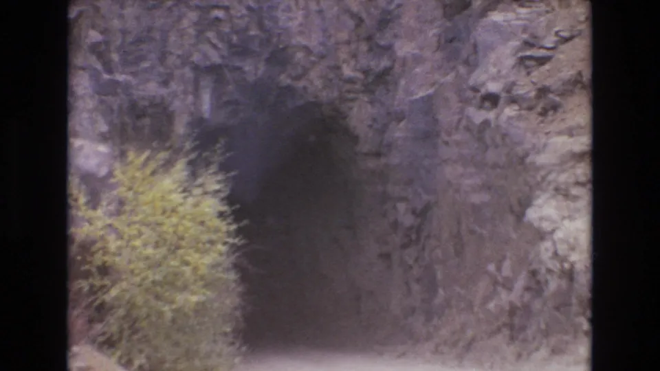 1973 Colorado Springs travel frame showing a dark tunnel opening carved into rock beside roadside brush and rugged stone.