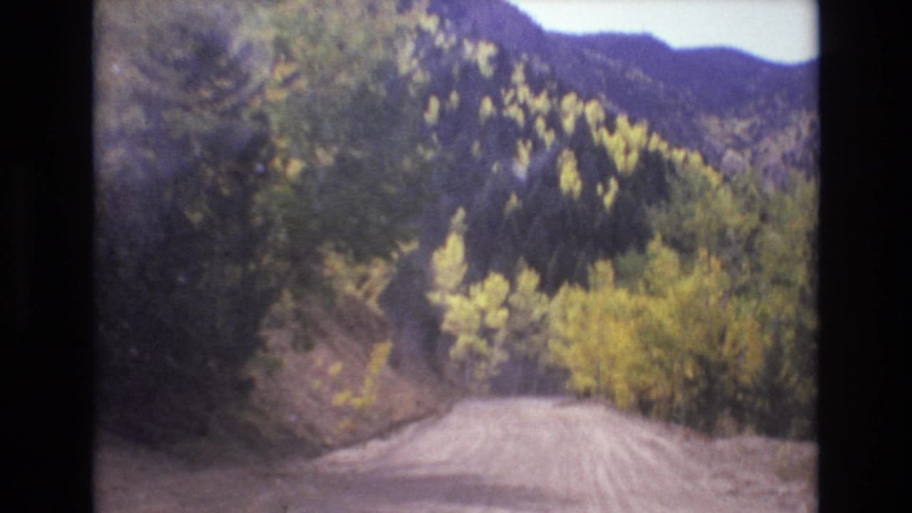 Dirt mountain road curving through forested slopes with yellow autumn trees.