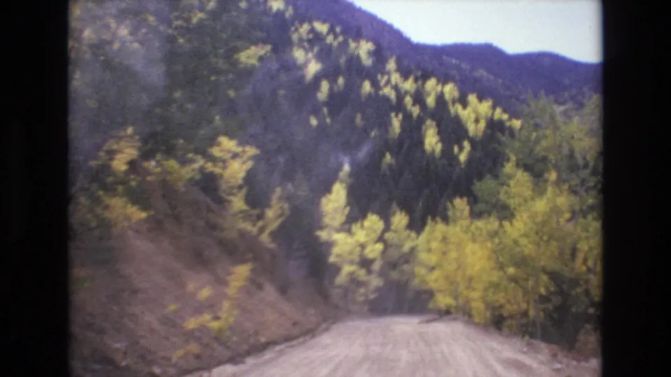 Forward-looking 8mm travel shot of an unpaved Colorado mountain road curving past steep banks, evergreens, and patches of yellow foliage.