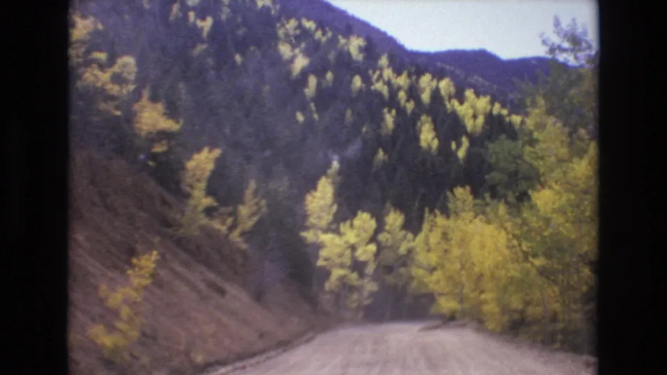 Passenger-view home-movie frame from the Colorado Springs area showing a dirt road descending through yellow autumn trees and dark wooded hills.