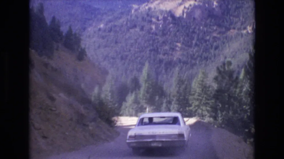 1973 8mm frame near Colorado Springs showing a white early 1970s American sedan ahead on a dusty mountain road bordered by evergreens and steep slopes.