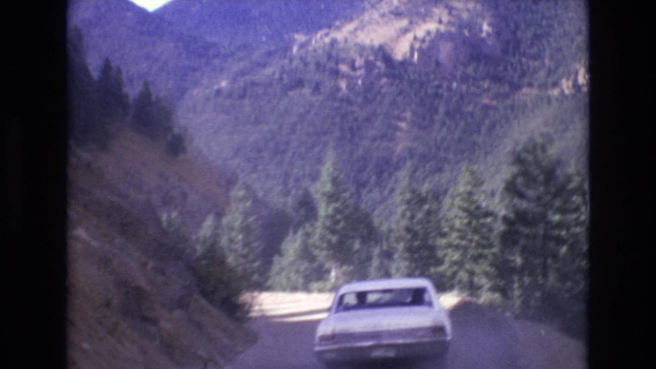 White sedan on a winding mountain road bordered by evergreen trees and steep slopes.
