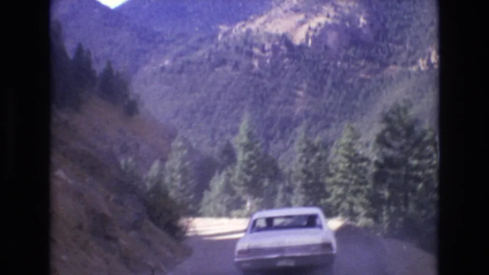 A white sedan drives away along a narrow mountain road near Colorado Springs, with evergreen pines, forested slopes, and distant ridges visible in 1973 home-movie footage.