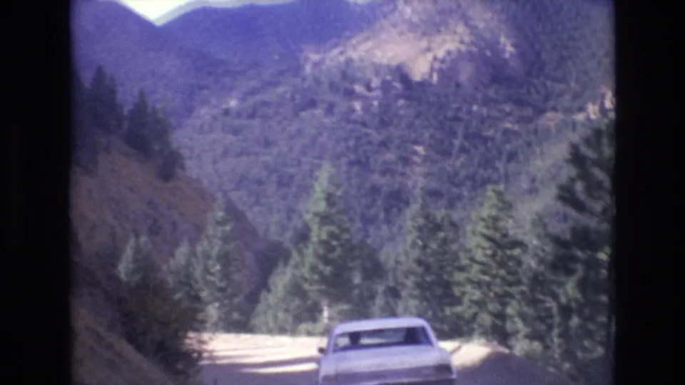 The white sedan moves through a bend on a wooded mountain road in the Colorado Springs area, with pine trees and rocky slopes filling the background.