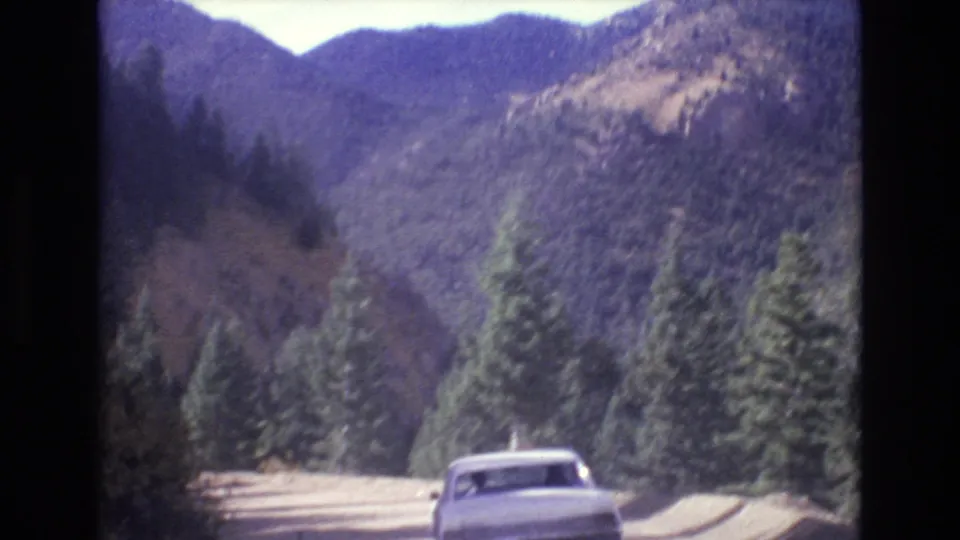 A rear view of the white sedan shows it traveling along a dirt mountain road, framed by tall evergreen trees and layered Colorado mountain terrain.