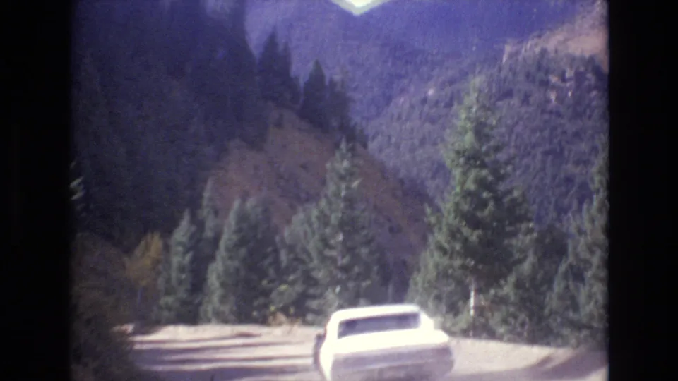 The white sedan recedes along the road between pine trees and shaded mountain slopes, with the soft color and exposure shifts of 1970s 8mm home-movie film.