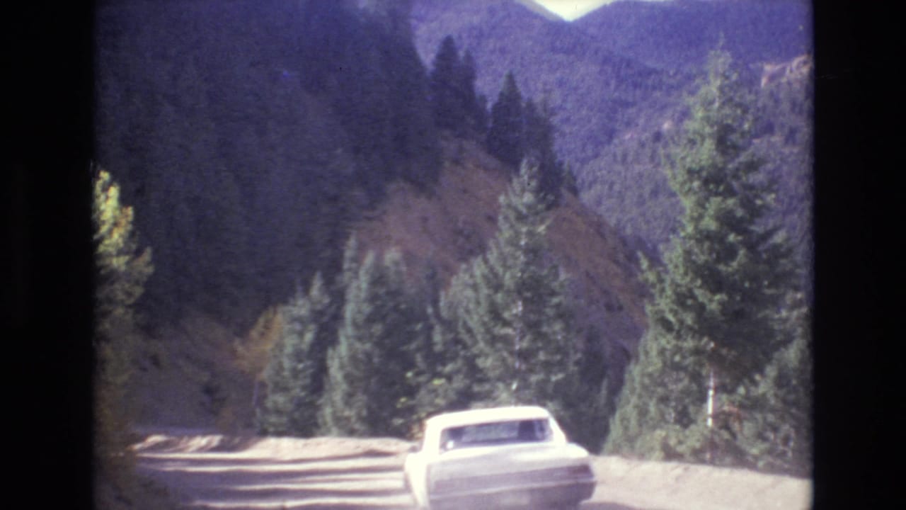 A white car drives ahead on a winding mountain road near Colorado Springs in metadata-supported 1973 footage, with evergreens and distant ridges visible.