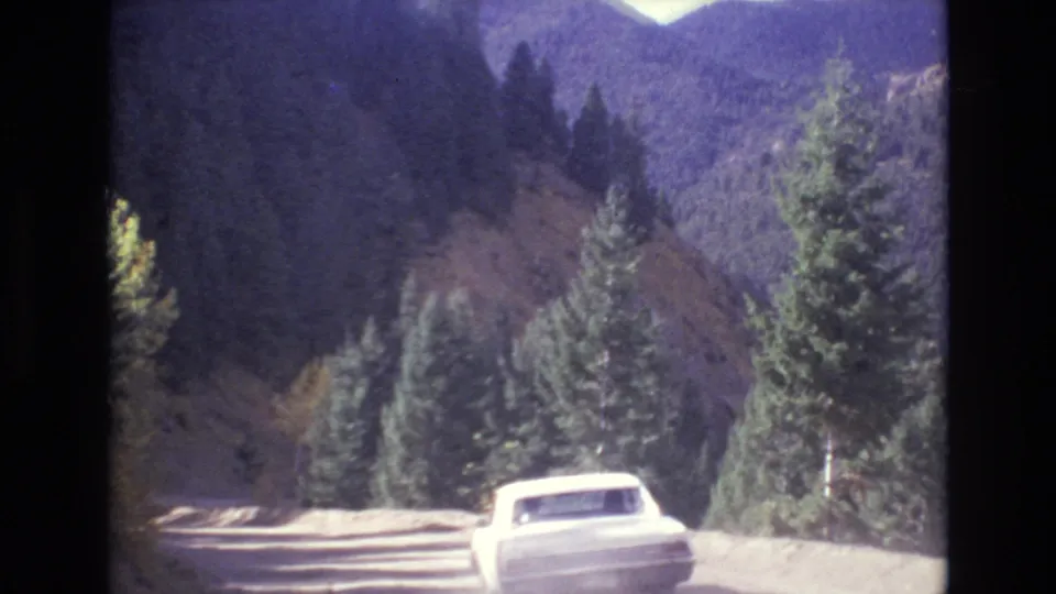 A white car drives ahead on a winding mountain road near Colorado Springs in metadata-supported 1973 footage, with evergreens and distant ridges visible.