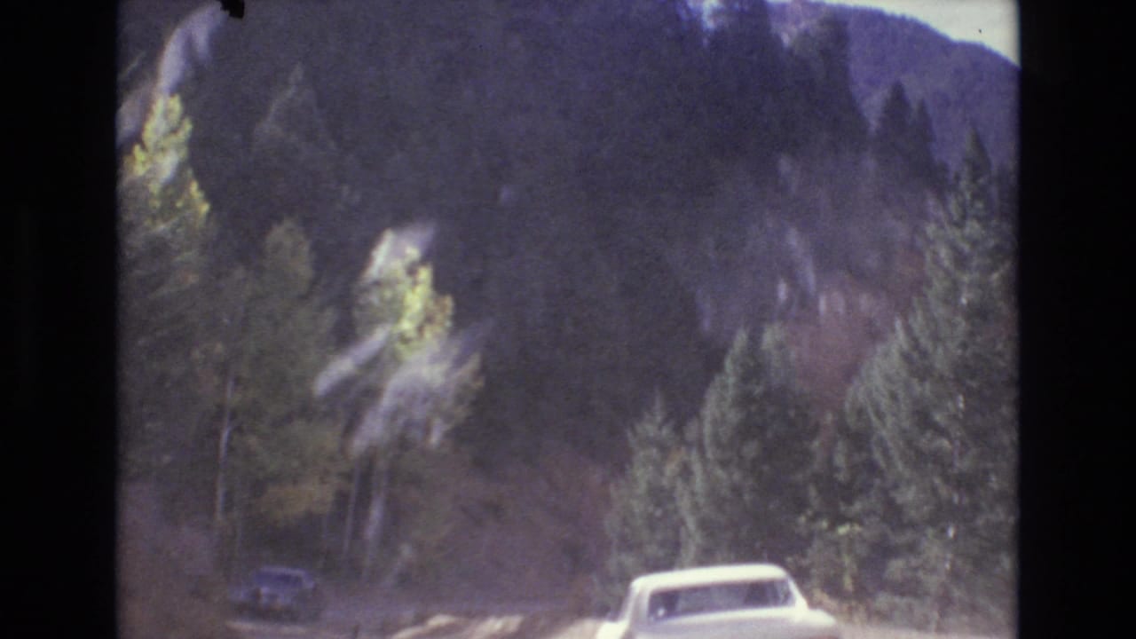 White car partly visible on a curving mountain road beneath dark forested hills.