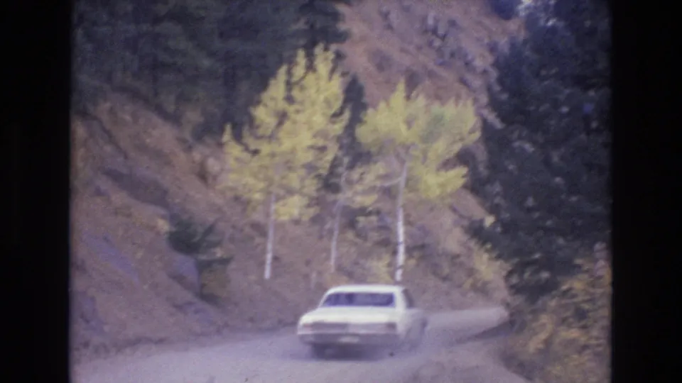 A white car drives past yellow autumn aspens and dark evergreens on a narrow mountain road in the 1973 Colorado Springs home-movie clip.