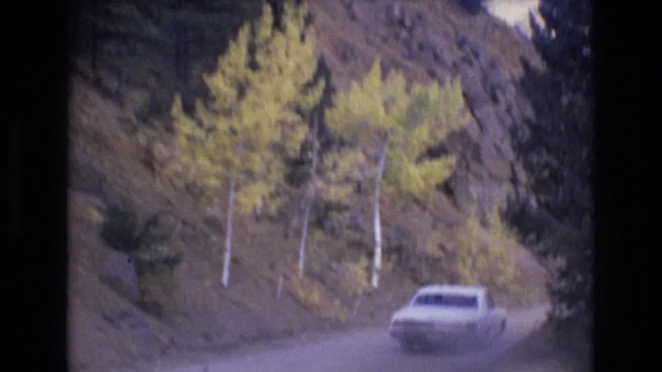 The white car recedes on the winding road beside yellow aspens, evergreen trees, and a rocky hillside in the Colorado Springs mountain setting.