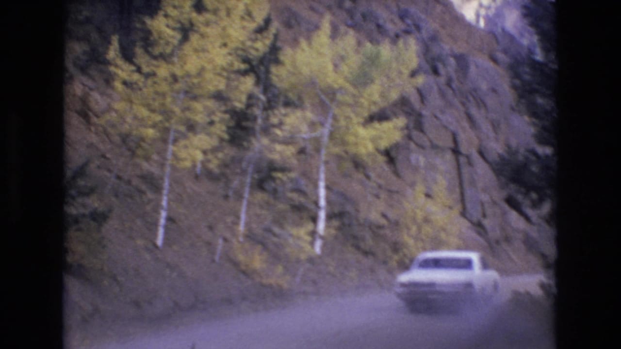 A white car drives along a dusty mountain road near Colorado Springs in 1973, passing rocky slopes and yellowing trees in soft 8mm color.