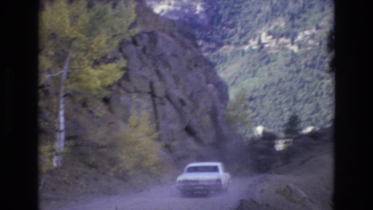The car continues along a narrow unpaved road in the Colorado Springs mountain landscape, with rock walls, trees, and a forested valley visible ahead.